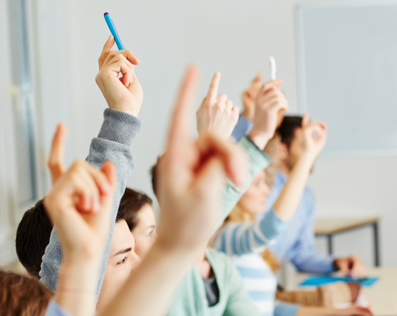 La imagen muestra estudiantes levantando la mano