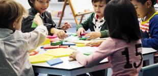 La imagen muestra un grupo de niños y niñas en una mesa interactuando con diversos objetos de tamaños y colores diferentes