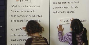La imagen muestra a dos niñas pegando palabras en un cuento en el pizarrón